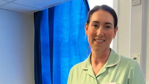 Clare Worden/BBC Jen Rudd is wearing a light green scrub dress. She had black hair tied back. She is standing in front of a hospital bed. 