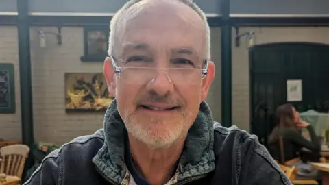 Andrew Sadler Andrew Sadler smiles at the camera as he is seated in a cafe. He is wearing a denim jumper and glasses. He has white hair and a white beard. 