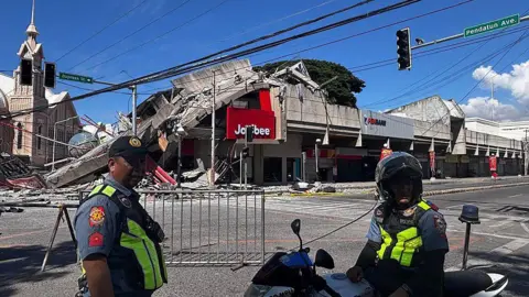 Policemen in uniform stand in front of a barricaded road. Behind them is a partially collapsed building with a red Jollibee sign.