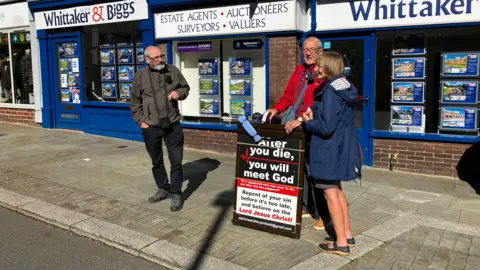 Two men and a woman on a street in Leek standing next to a sign, saying: "After you die you will meet God - Repent of your sin before it's too late, and believe on the Lord Jesus Christ!"  