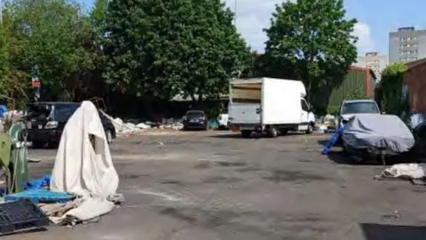 A disused car park with various vehicles parked randomly around the site. A white van is on the far side and rubbish is strewn around the edges.