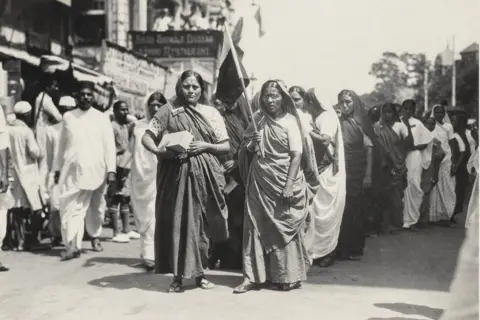 The Alkazi Collection of Photography Two women in saris and with their heads covered, are seen leading a boycott procession in India. The photo, which is monochrome, shows them walking along a busy street. The woman on the right is wearing glasses and is holding a provisional national flag. 
