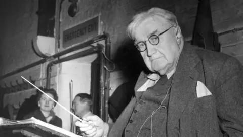 Getty Images A man wearing a suit and glasses holding a conductor's baton.
