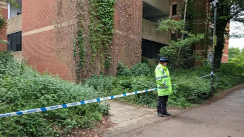 A female police officer wearing a hi-vis coat, black hat, black trousers and black shoes stands in front of a cordon that has been erected in front of a multi-storey car park. The officer is looking away from the camera.