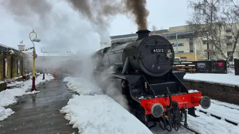 BBC Weather Watchers/Chris King A steam train sits in a station with snow on the platforms. Black smoke comes out of its chimney and steam from underneath it.