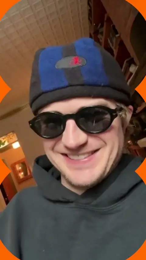 A man, Djo, smiles wearing a beanie hat, sunglasses and hoodie