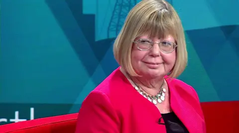 Head and shoulder shot of Liz Twist who has blonde bobbed hair. She is wearing glasses, a red jacket, with a dark dress or blouse underneath, and a necklace featuring small white disks. The back of a red sofa can be seen behind her, against a background in shades of blue.