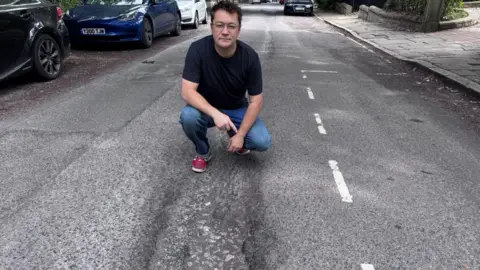 Molly Rusling A man has black hair, glasses, a black T-shirt, jeans and red trainers. He is crouched down in the middle of a road next to a very long and wide pothole.