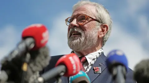 PA Media Gerry Adams outside the High Court in Dublin, after he won his libel action against the BBC. He is smiling and speaking into multiple microphones. He has grey hair and is wearing glasses. He is wearing a blue suit and red patterned tie.