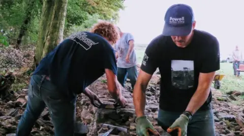 Three men are building a drystone wall. The one in the foreground is wearing gloves and placing a stone on top of the wall. The man opposite him is also placing a stone on top of the waist-high wall. There are spades and a wheelbarrow behind them.