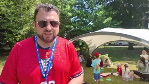 Gary Spracklen, headteacher at The Prince of Wales School, giving interview while the children are playing in the shade behind him.
