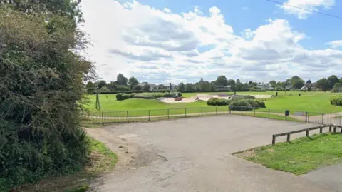 A Google street view image of the entrance to a large green park with a skate park, including ramps, in the centre of the frame.