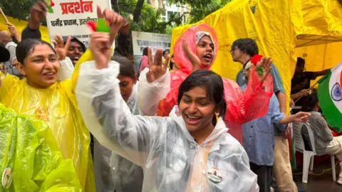 Geeta Pandey A girl wearing a raincoat is carrying a plastic rose. She is surrounded by other protesters raincoats.