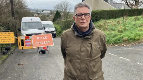 Bob Hembry, who has his hand behind his back, is wearing a green jacket and glasses. He is standing in front of a grass verge and a sign saying "road closed".