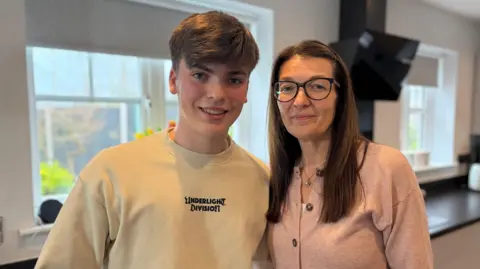 BBC A young man with short dark hair in a cream sweatshirt next to his mother with long dark hair, glasses and a pink cardigan. They are standing in a kitchen
