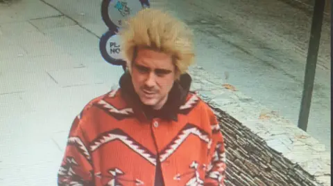 Alexander Key is walking in a street. He has long peroxided hair standing up and a moustache. He is wearing a bright red coat which has a bold zigzag print in white and black.