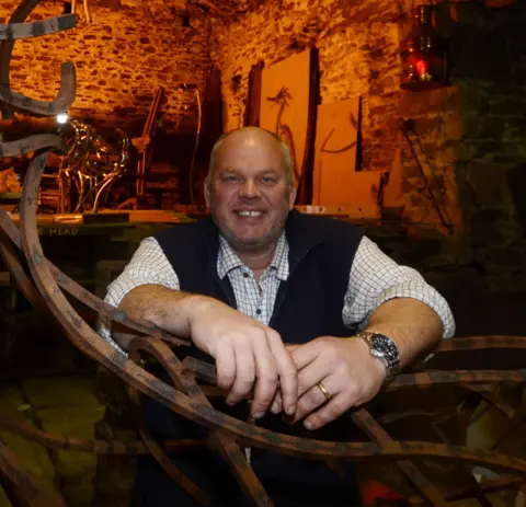 Andy Kay Andy Kay leaning on one of his steel sculptures inside his workshop. He has a bald head with short grey hair at the sides. He is wearing a check shirt with the sleeves rolled up, with a black waistcoat over the top. He is smiling. The stone inner wall of his workshop is bathed in an orange glow behind him.