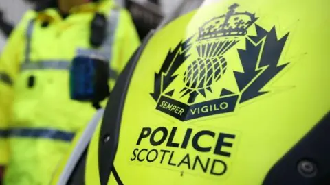 A yellow police vehicle with Police Scotland wording, and an officer in the background.