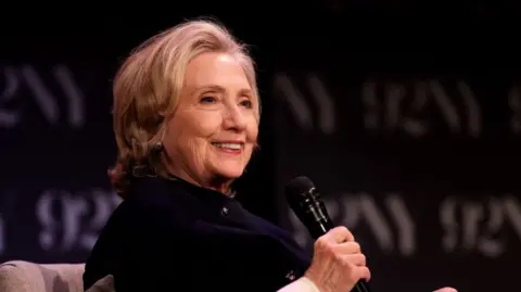 Hillary Clinton has short blonde hair which is curled in at the bottom. She has a black top on and is wearing makeup as she holds a black microphone.