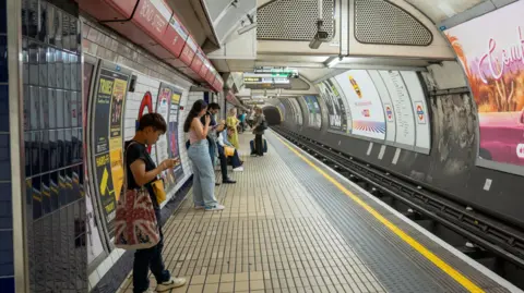 AFP via Getty Images People standing on a Tube platform, which is adorned with poster adverts