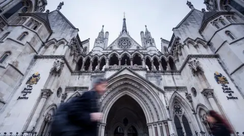 An exterior view of the Royal Courts of Justice, taken from a low viewpoint, showing the neo-Gothic arches and spires reaching towards the sky as figures blurred by their movement pass in front of the building