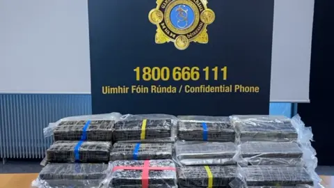 An Garda Síochána Bundles of suspected cocaine in front of a blue Gardai logo with a confidential telephone number in yellow