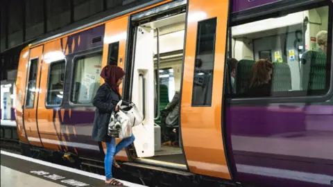 A woman wearing a red hijab, blue coat and blue jeans is walking onto an orange and purple train carriage while holding bags.