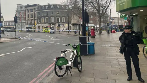 A police officer stands in uniform to the right on the pavement of Lewisham High Street with a Lime bike in the foreground. In the background there are two banks - Barclays sign in view, with police tape blocking the road.