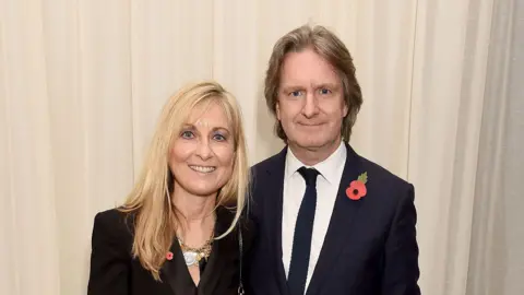 Getty Images Fiona Phillips and Martin Frizell stand against a beige background. The photo is landscape - both smile and look towards the camera.