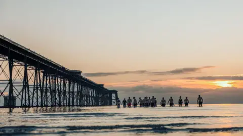 Visit Isle of Man A group of swimmers in trunks and swimming costumes walking out of the sea at Ramsey at sunrise, with the water level is up to their knees. The Queen's Pier, which is a long iron pier, can be seen in the background in silhouette. The sky has pink hues.