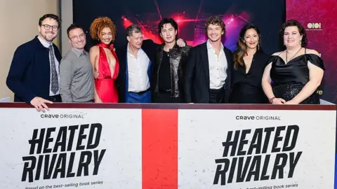 Getty Images Brendan Brady, Jacob Tierney, Ksenia Daniela Kharlamova, Dylan Walsh, Hudson Williams, Connor Storrie, Christina Chang and Rachel Reid stand in a line behind a Heated Rivalry billboard the Toronto premiere 