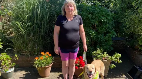 Jenny Stafford Jenny Stafford is smiling at the camera and her guide dog Nan. She is wearing a black running top with pink shorts. In the background is greenery in a garden.