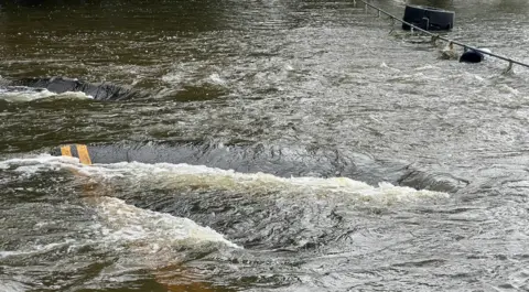 Harriet Heywood/BBC An image of Mill Lane fully submerged under water. There is a chunk of tarmac with double yellow lines on sticking out of the water.