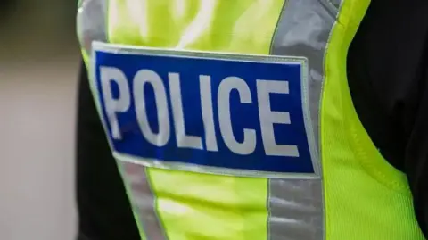 A police officer (photographed from behind) wearing a yellow high visibility jacket which has 'Police' written on it in white lettering on a blue background. 