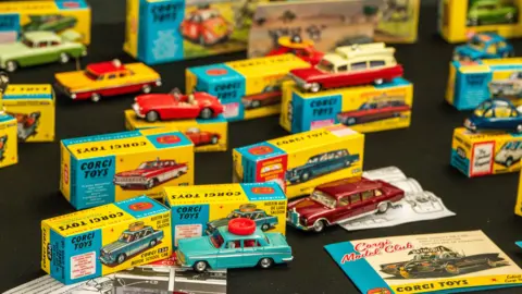 A collection of mid‑20th‑century Corgi Toys model cars with their colourful yellow‑and‑blue boxes behind them. Leaflets and instruction sheets from the Corgi Model Club are scattered among the toys.