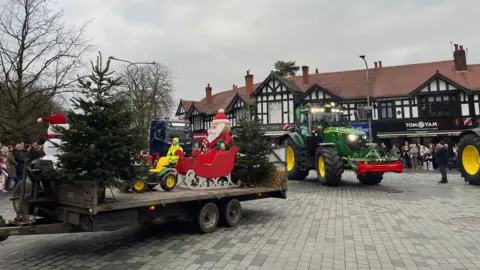 BBC/ Zoe Toase A Santa Christmas display on a cart pulled by a tractor in a town square, with other tractors following behind 