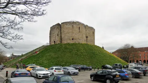 Google A large, circular stone tower sitting on top of a steep grassy hill. The tower looks old and historic, built from pale stone blocks, with narrow vertical slits that resemble medieval arrow loops. A long staircase runs diagonally up the left side of the hill, with a few people walking along it. At the bottom of the hill, there is a car park filled with various modern cars.