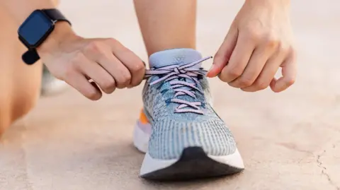 Close-up of hands tying the laces on a blue running shoe worn on a paved surface. The person is crouched down, wearing light-colored socks and a smartwatch on one wrist, with the focus on the shoelaces being pulled tight.
