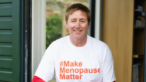 Jenny Smith photography Diane Danzebrink⁢ in a doorway smiling at the camera. She⁤ wears a white‍ T-shirt with the words⁣ '#Make Menopause Matter' written ⁤in orange letters in the center. 