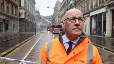 PA Media Swinney wearing a bright orange ScotRail high‑visibility jacket stands in front of an emergency cordon on a wet city street, with fire crews and fire engines working in the background amid smoke