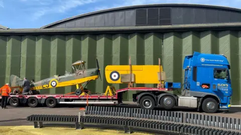 Bottisham Airfield Museum Tiger Moth N6635 on a lorry trailer in front of a hangar at the Imperial War Museum, Duxford. t has a grey body with a bright yellow underbelly. Its two wings have been removed and are resting on the trailer in front of its nose. The lorry cab is bright blue.