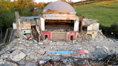 Owen McNutt Edendork Hall with a lot of rubble on the floor and a frame of the building with the back wall still intact. The dome is also still up which is grey and has small specks of gold left on it. Behind the demolished building is a green field. The image has been taken during sunset so the grey building is reflected by a reddish sunlight. 