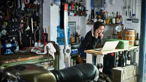 Getty Images Female mechanic sitting in a workshop and calling someone on the phone while looking at a laptop computer