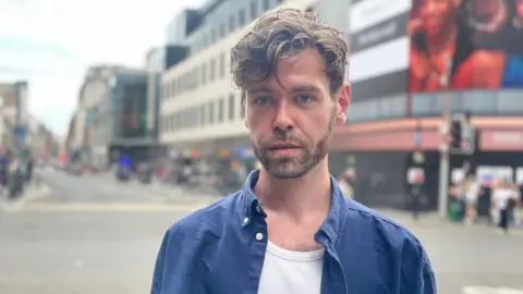 Portrait of Kip Lawson standing on a city street, wearing a blue button-up shirt over a white vest. The background is softly blurred, showing buildings, shopfronts, pedestrians, and a road intersection.