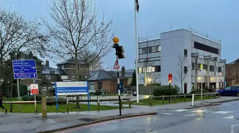 An entrance to Watford General Hospital, showing a Watford General Hospital sign in white lettering on a blue background. Another sign gives directions for various car parks. There is a three-storey white building to the right, with black window frames and some windows lit up. There is a two-storey brick building in the background, and a road runs past in the foreground.