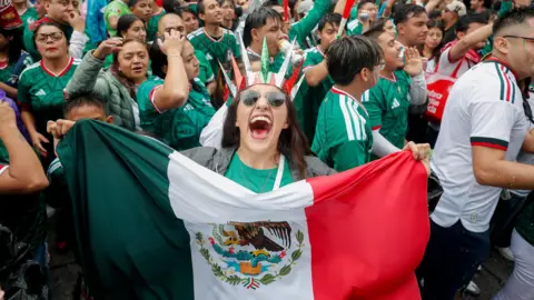 A person wearing green holds a Meican flag to the camera, with a crowd of people celebrating visible behind her.