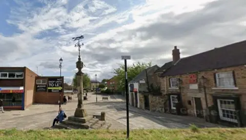 The pedestrianised Market Square in Woodhouse, Sheffield with an old market stone cross/war memorial in the centre. A man in blue with a rucksack is sitting on the steps of the cross. An old stone building, a pub, is to the right of the picture with shops to the left. A hedge is in the foreground of the picture.