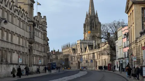 EstherJ An Oxford street is busy with shoppers. Stone buildings line each side of the road and there is a blue bus. It is a sunny day. In the centre of the picture is a building with a tall stone spire.