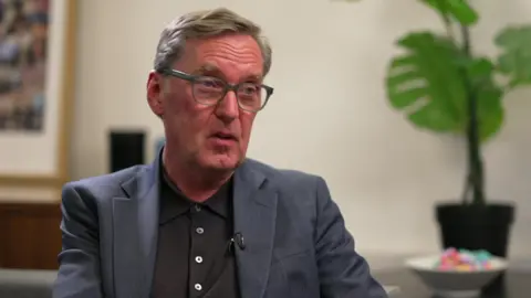 Alan Milburn pictured wearing glasses, a blazer and black polo t-shirt. 