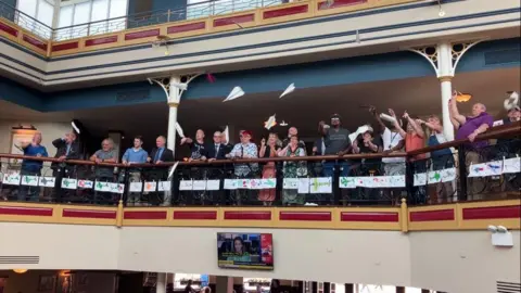 A group of people are standing on a balcony and can be seen launching paper aeroplanes into the air.